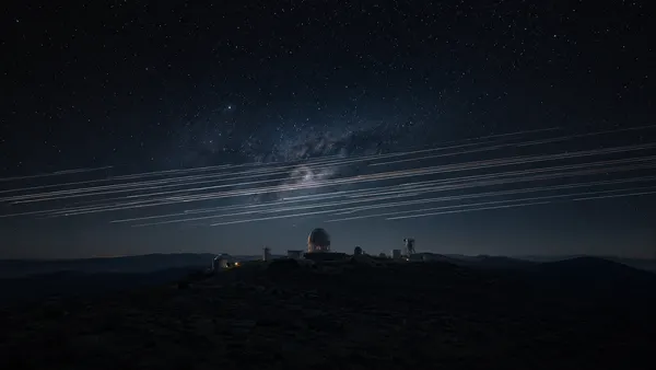 Bright Starlink satellite trails crossing the night sky above a remote astronomical observatory, with the Milky Way arcing faintly behind